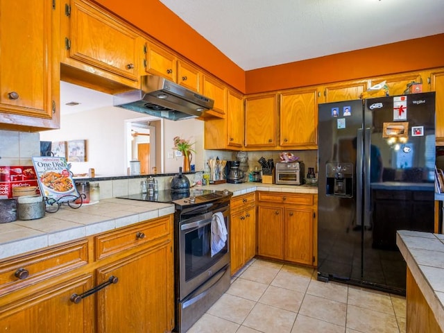 kitchen featuring light tile patterned floors, brown cabinetry, stainless steel electric range oven, under cabinet range hood, and black fridge