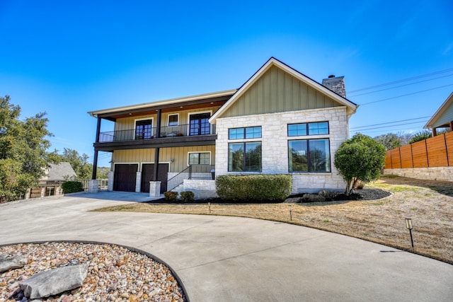 view of front facade featuring concrete driveway, a balcony, stone siding, a chimney, and fence