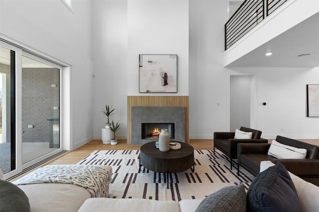 living room with a lit fireplace, light wood-type flooring, a towering ceiling, and baseboards