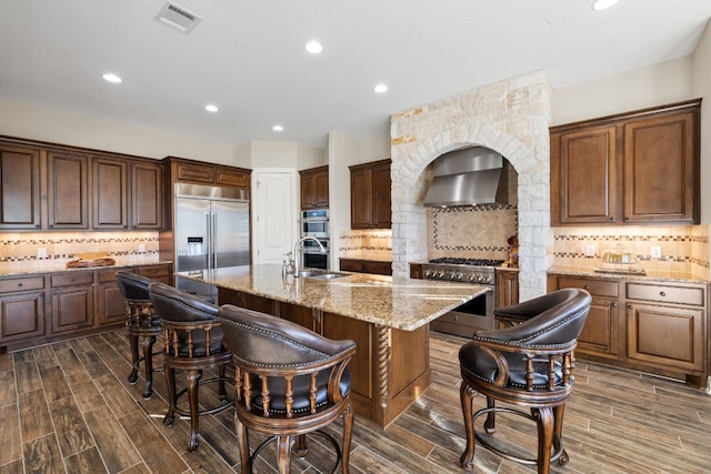 kitchen featuring a breakfast bar, high end appliances, visible vents, a kitchen island with sink, and wall chimney range hood