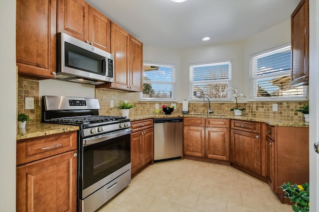 kitchen featuring light stone counters, stainless steel appliances, backsplash, brown cabinetry, and a sink