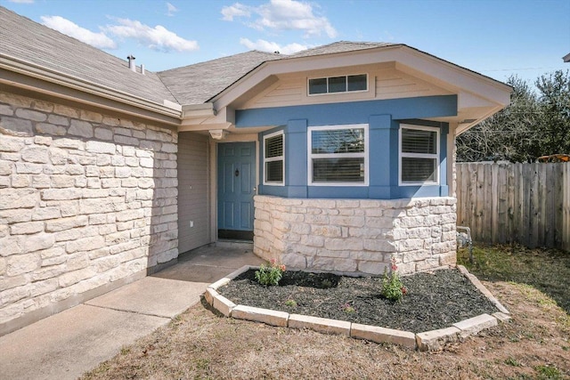 view of exterior entry featuring stone siding, a shingled roof, and fence