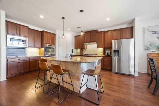 kitchen with under cabinet range hood, stainless steel appliances, dark wood-style flooring, tasteful backsplash, and a kitchen bar