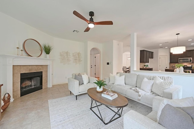 living area featuring light tile patterned floors, visible vents, arched walkways, a ceiling fan, and a tiled fireplace