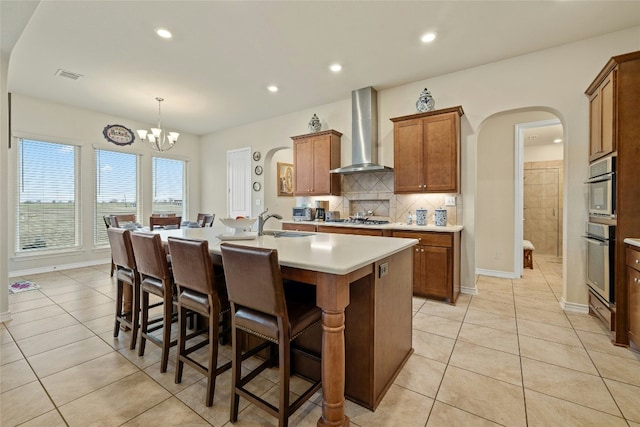 kitchen with arched walkways, wall chimney exhaust hood, appliances with stainless steel finishes, brown cabinets, and a kitchen island with sink