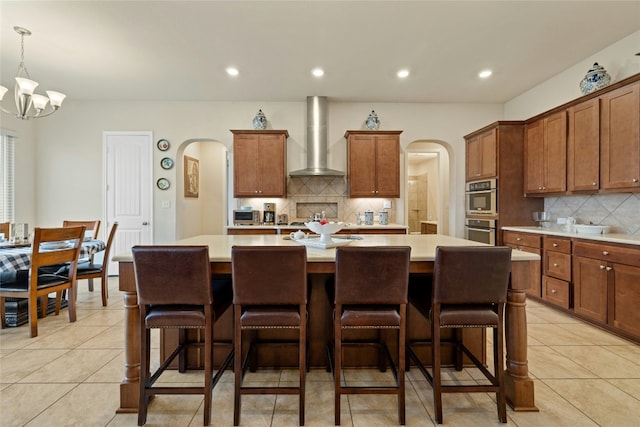 kitchen with arched walkways, light countertops, wall chimney range hood, and a kitchen island with sink