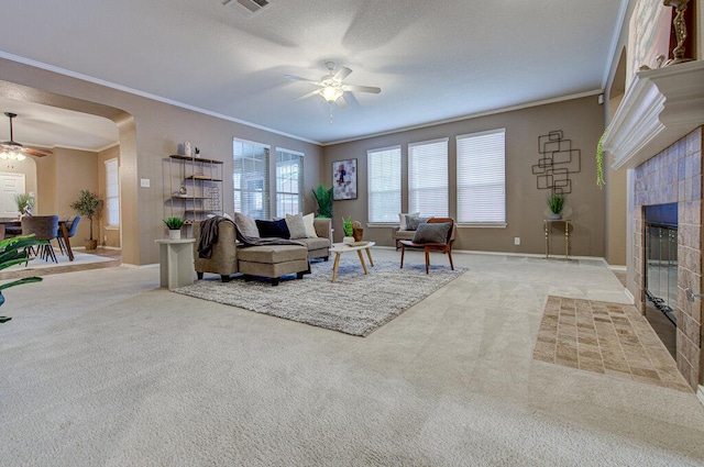 living area featuring arched walkways, a ceiling fan, a tiled fireplace, ornamental molding, and carpet