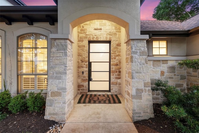 exterior entry at dusk with stone siding, a shingled roof, and stucco siding