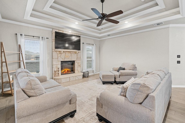 living room featuring a stone fireplace, wood finished floors, visible vents, a ceiling fan, and a tray ceiling