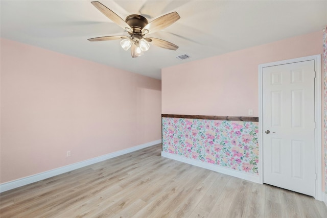 empty room with light wood-style flooring, a wainscoted wall, a ceiling fan, visible vents, and baseboards