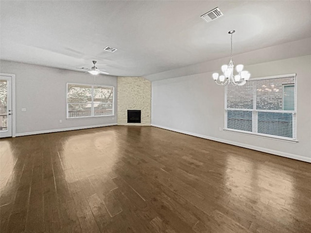 unfurnished living room featuring a stone fireplace, ceiling fan with notable chandelier, dark wood-style flooring, visible vents, and baseboards