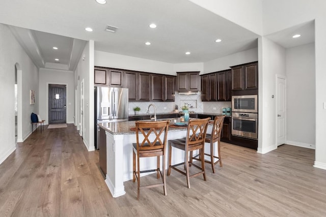 kitchen featuring light stone counters, dark brown cabinets, appliances with stainless steel finishes, tasteful backsplash, and an island with sink