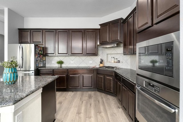 kitchen featuring stainless steel appliances, decorative backsplash, dark brown cabinetry, dark stone countertops, and light wood-type flooring