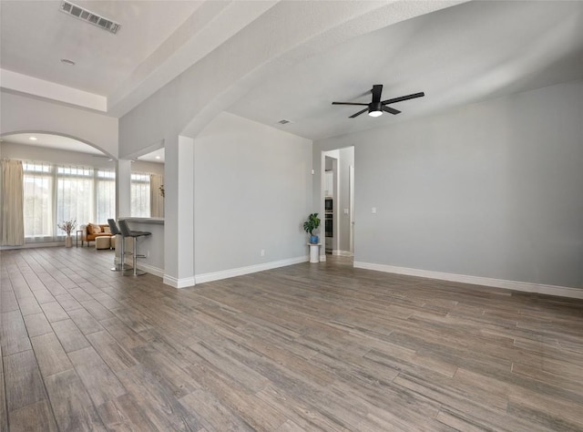 unfurnished living room with arched walkways, ceiling fan, wood finished floors, visible vents, and baseboards