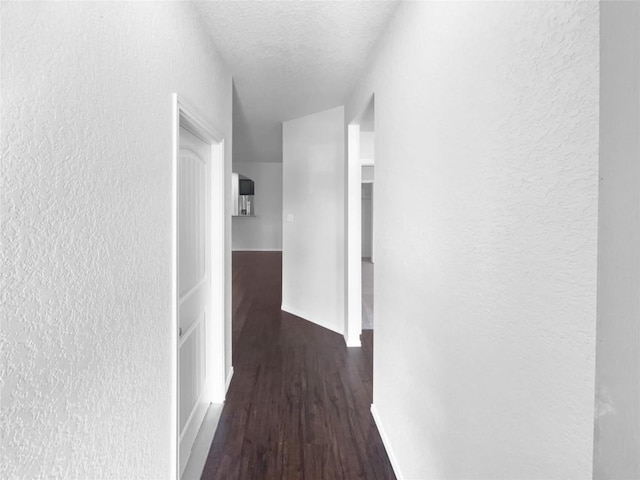 hallway with a textured ceiling, a textured wall, wood finished floors, and baseboards