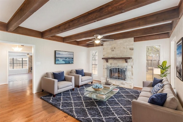 living area featuring a ceiling fan, beamed ceiling, a stone fireplace, and wood finished floors