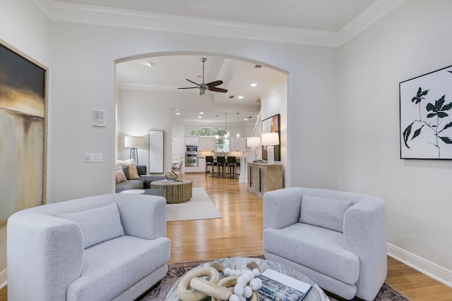 living area featuring light wood finished floors, baseboards, arched walkways, a ceiling fan, and crown molding