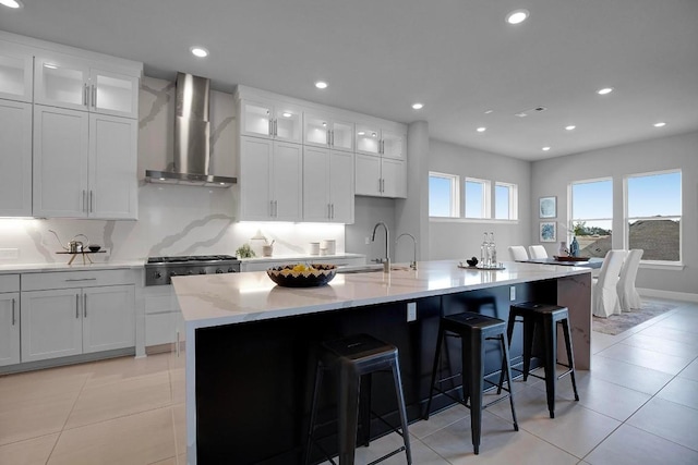 kitchen with a sink, white cabinetry, a kitchen island with sink, and wall chimney range hood