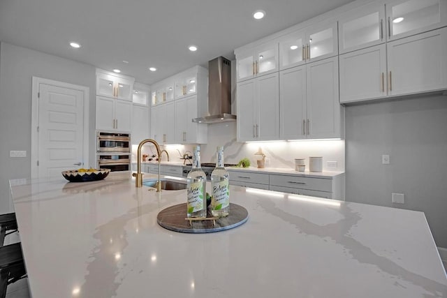 kitchen with light stone countertops, stainless steel double oven, white cabinets, wall chimney exhaust hood, and a sink