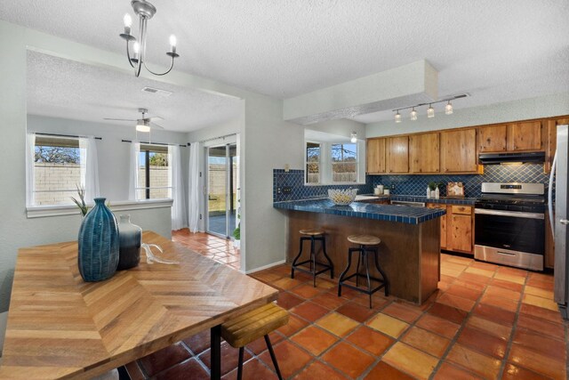 kitchen with under cabinet range hood, stainless steel appliances, backsplash, brown cabinetry, and dark countertops