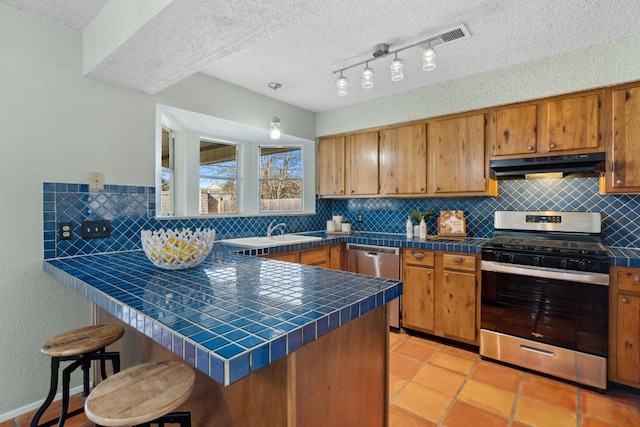 kitchen with tile counters, brown cabinetry, appliances with stainless steel finishes, a peninsula, and under cabinet range hood