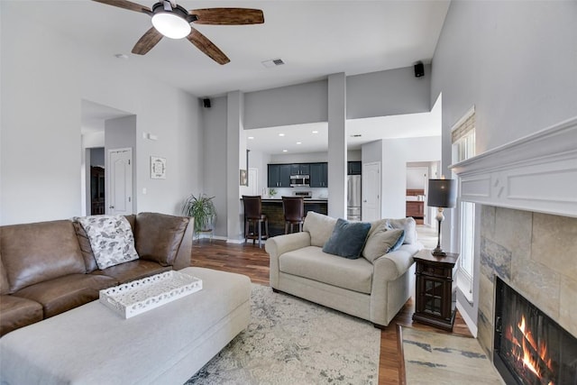 living room featuring a towering ceiling, ceiling fan, wood finished floors, a tile fireplace, and baseboards