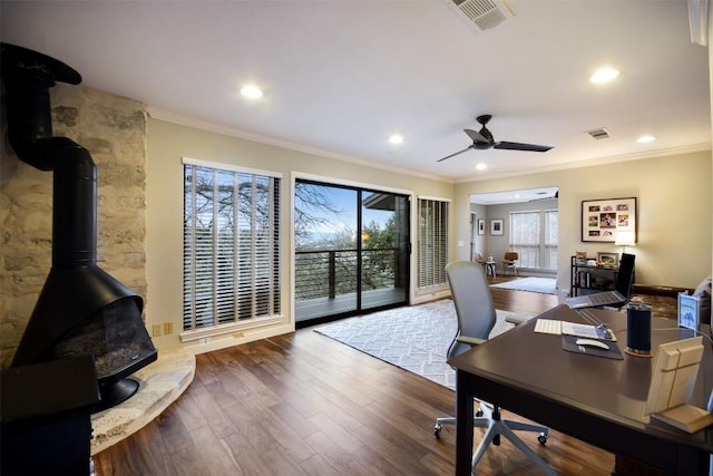 home office featuring crown molding, visible vents, a wood stove, ceiling fan, and wood finished floors