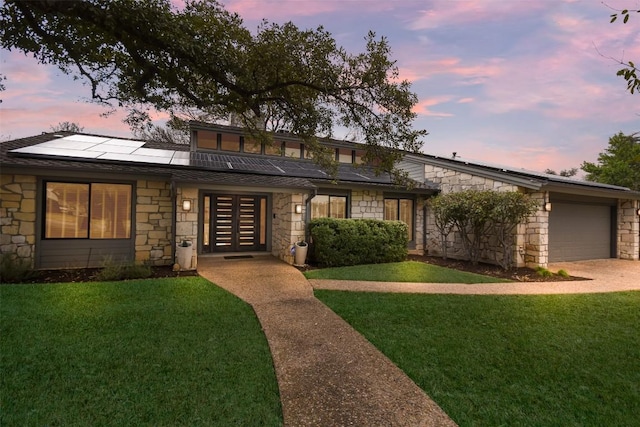 mid-century home featuring a garage, stone siding, roof mounted solar panels, and a lawn