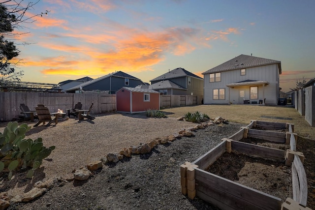 yard at dusk with an outbuilding, a vegetable garden, a storage unit, a fenced backyard, and a fire pit