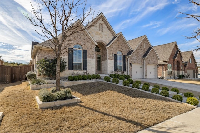 french country inspired facade with a garage, concrete driveway, stone siding, fence, and brick siding