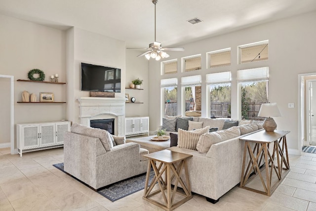 living area with a high ceiling, a ceiling fan, visible vents, baseboards, and a glass covered fireplace