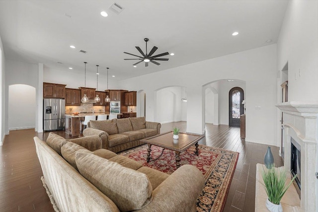 living area featuring arched walkways, a fireplace with raised hearth, visible vents, a ceiling fan, and dark wood-style floors