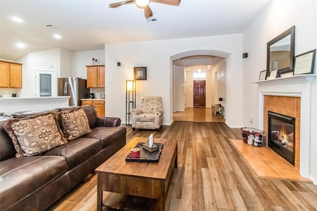 living room with arched walkways, ceiling fan, visible vents, light wood-style floors, and a tiled fireplace