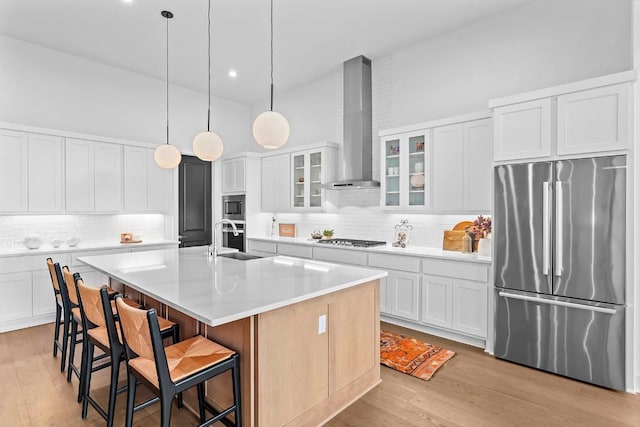 kitchen featuring a kitchen island with sink, light wood-style flooring, stainless steel appliances, decorative backsplash, and wall chimney exhaust hood