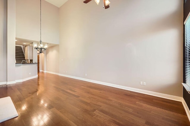 unfurnished living room with dark wood-type flooring, a towering ceiling, baseboards, and ceiling fan with notable chandelier