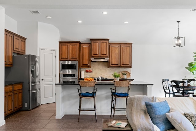 kitchen featuring dark countertops, appliances with stainless steel finishes, under cabinet range hood, a kitchen bar, and backsplash