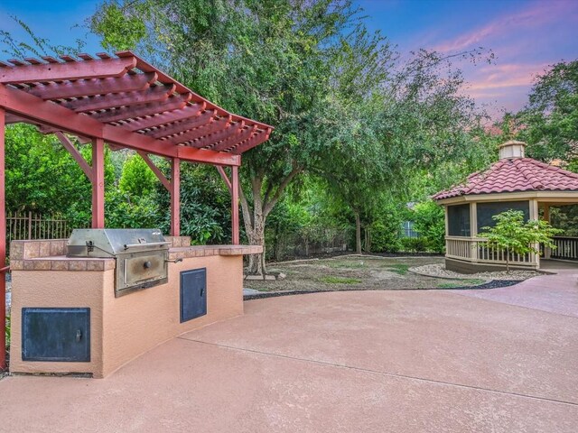 patio terrace at dusk featuring fence, exterior kitchen, a gazebo, area for grilling, and a pergola