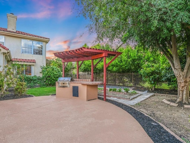 patio terrace at dusk with a grill, fence, a pergola, and an outdoor kitchen