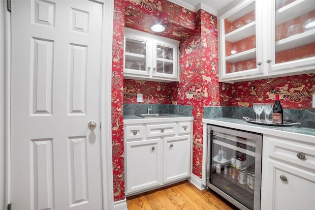 kitchen featuring wallpapered walls, light wood finished floors, wine cooler, white cabinetry, and a sink