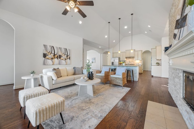 living room featuring arched walkways, high vaulted ceiling, a stone fireplace, hardwood / wood-style flooring, and a ceiling fan