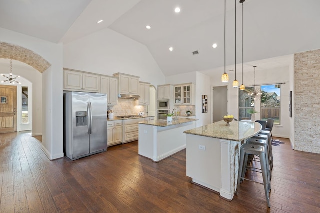 kitchen with arched walkways, stainless steel appliances, cream cabinets, an inviting chandelier, and an island with sink