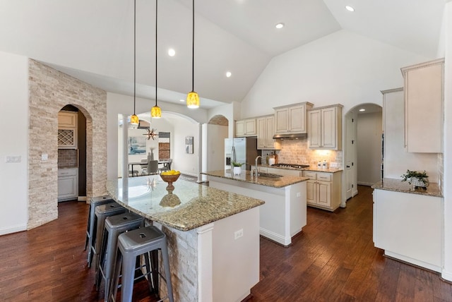 kitchen with dark wood-style floors, arched walkways, cream cabinetry, tasteful backsplash, and a kitchen island with sink