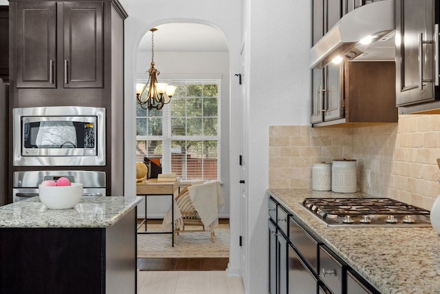 kitchen featuring arched walkways, dark brown cabinetry, under cabinet range hood, stainless steel appliances, and tasteful backsplash