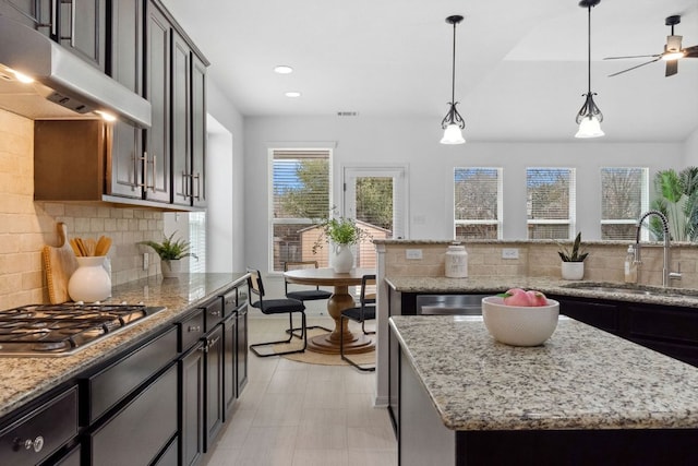 kitchen featuring under cabinet range hood, stainless steel gas cooktop, a kitchen island, a sink, and light stone countertops