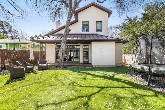 back of property with a trampoline, a chimney, stucco siding, a standing seam roof, and fence