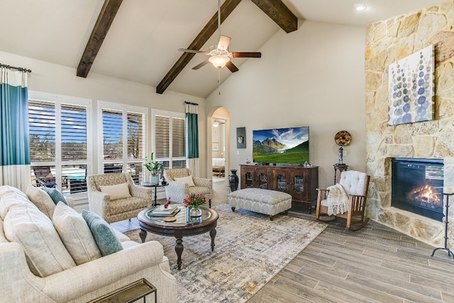 living area featuring arched walkways, wood finished floors, a stone fireplace, high vaulted ceiling, and beam ceiling