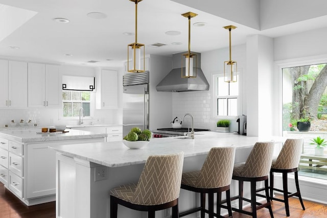 kitchen with wall chimney range hood, white cabinets, a sink, and built in fridge