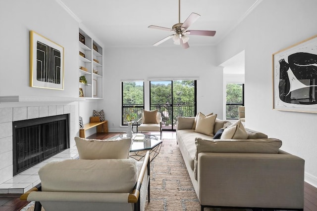 living room featuring wood finished floors, a ceiling fan, baseboards, ornamental molding, and a tiled fireplace