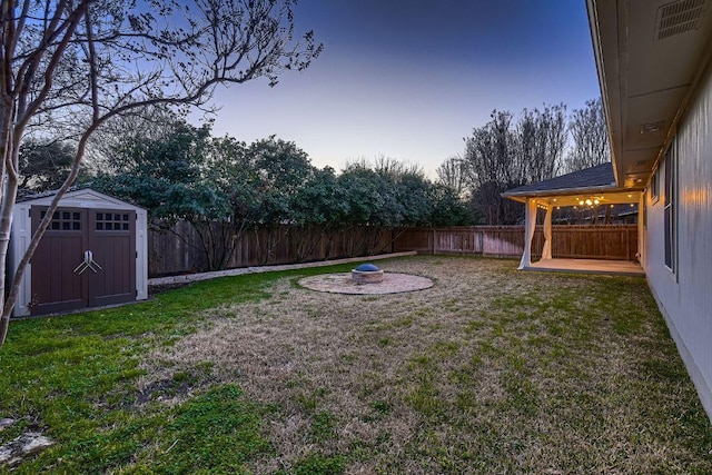 yard at dusk featuring a storage shed, a fire pit, a patio, a fenced backyard, and an outbuilding