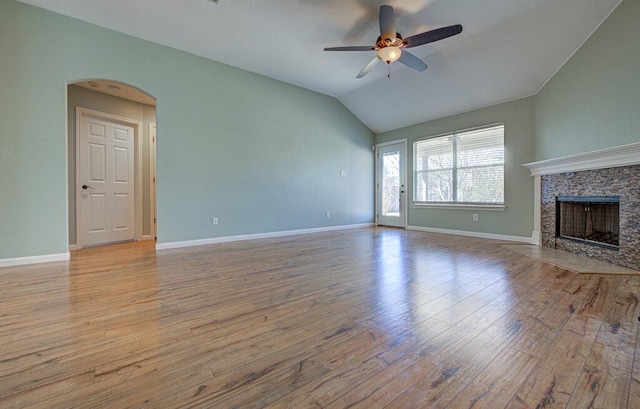 unfurnished living room featuring arched walkways, ceiling fan, a stone fireplace, hardwood / wood-style flooring, and vaulted ceiling